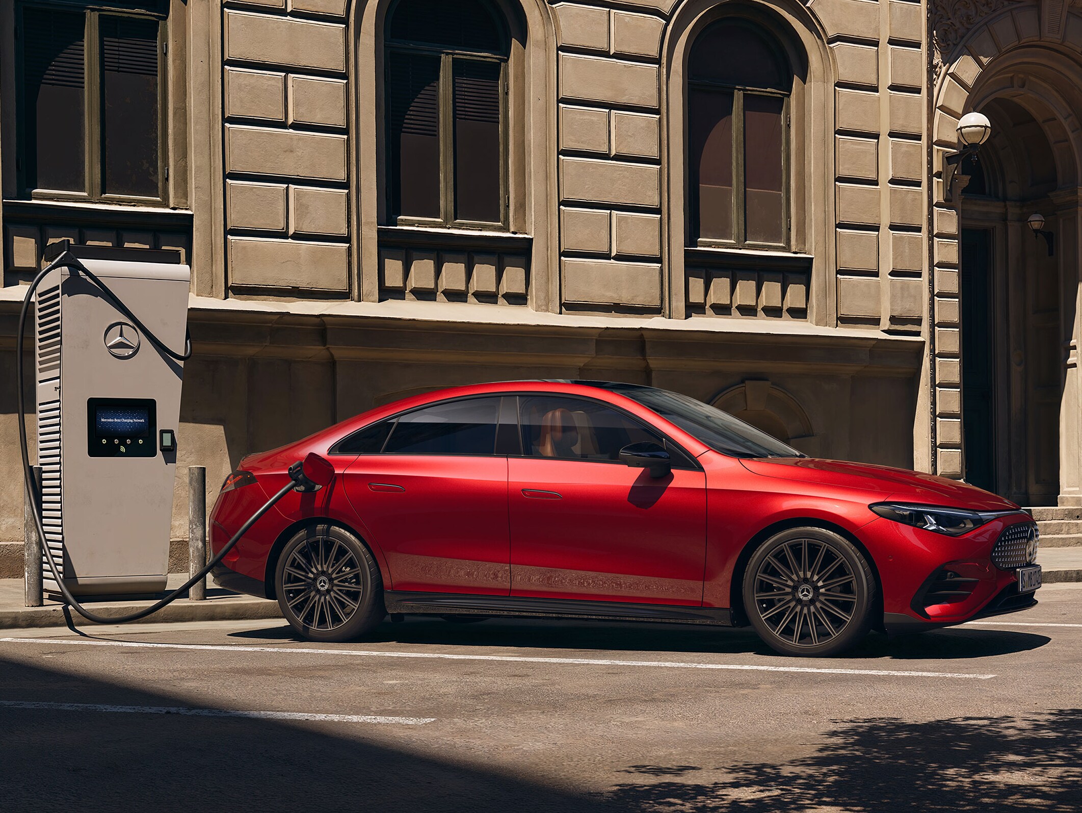 Röd Mercedes-Benz CLA Electric ansluten till en laddstation, parkerad vid trottoarkanten.