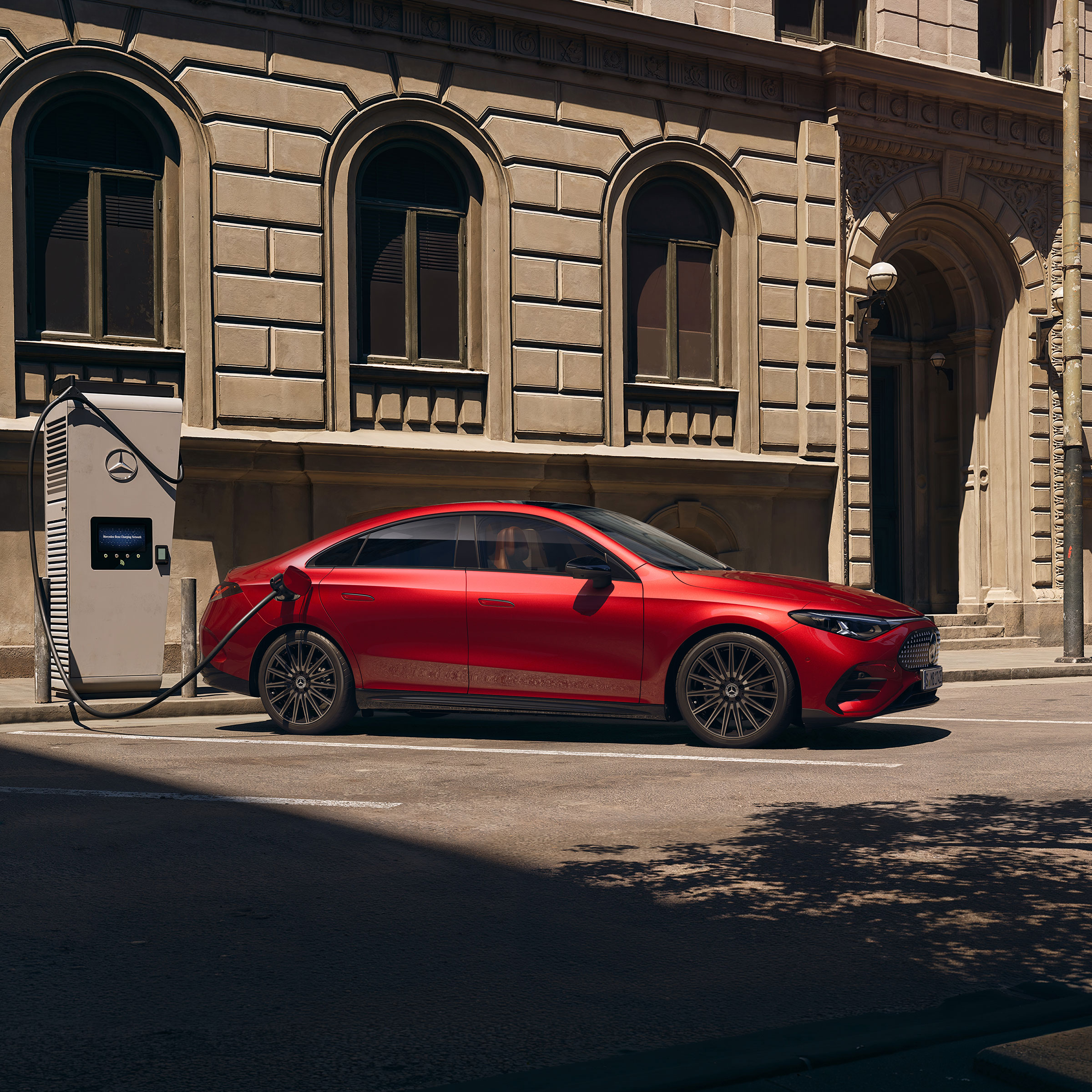 CLA electric i Online Store | Aktuella erbjudanden | Mercedes-Benz Röd Mercedes-Benz CLA Electric ansluten till en laddstation, parkerad vid trottoarkanten.