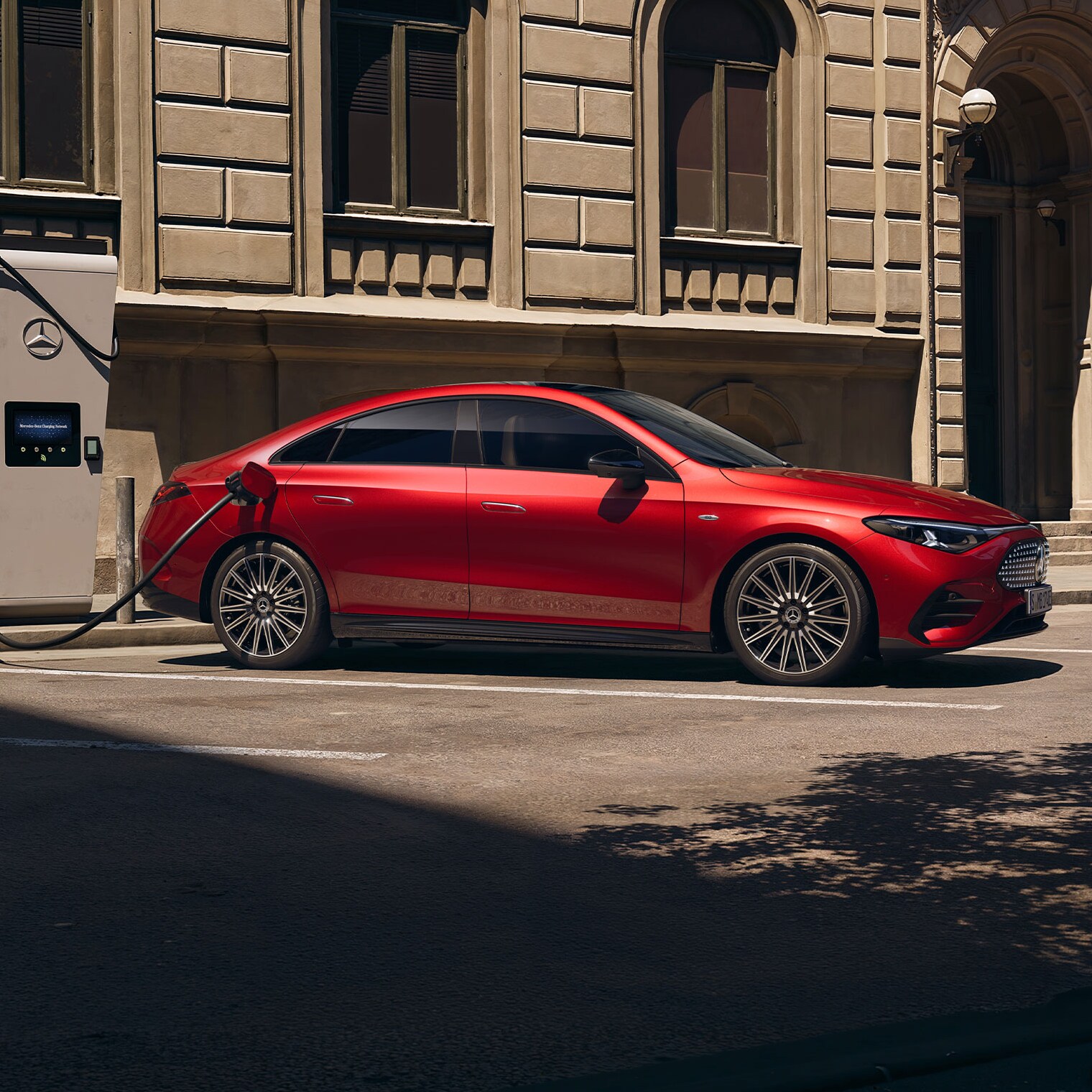 Röd Mercedes-Benz CLA Electric ansluten till en laddstation, parkerad vid trottoarkanten.