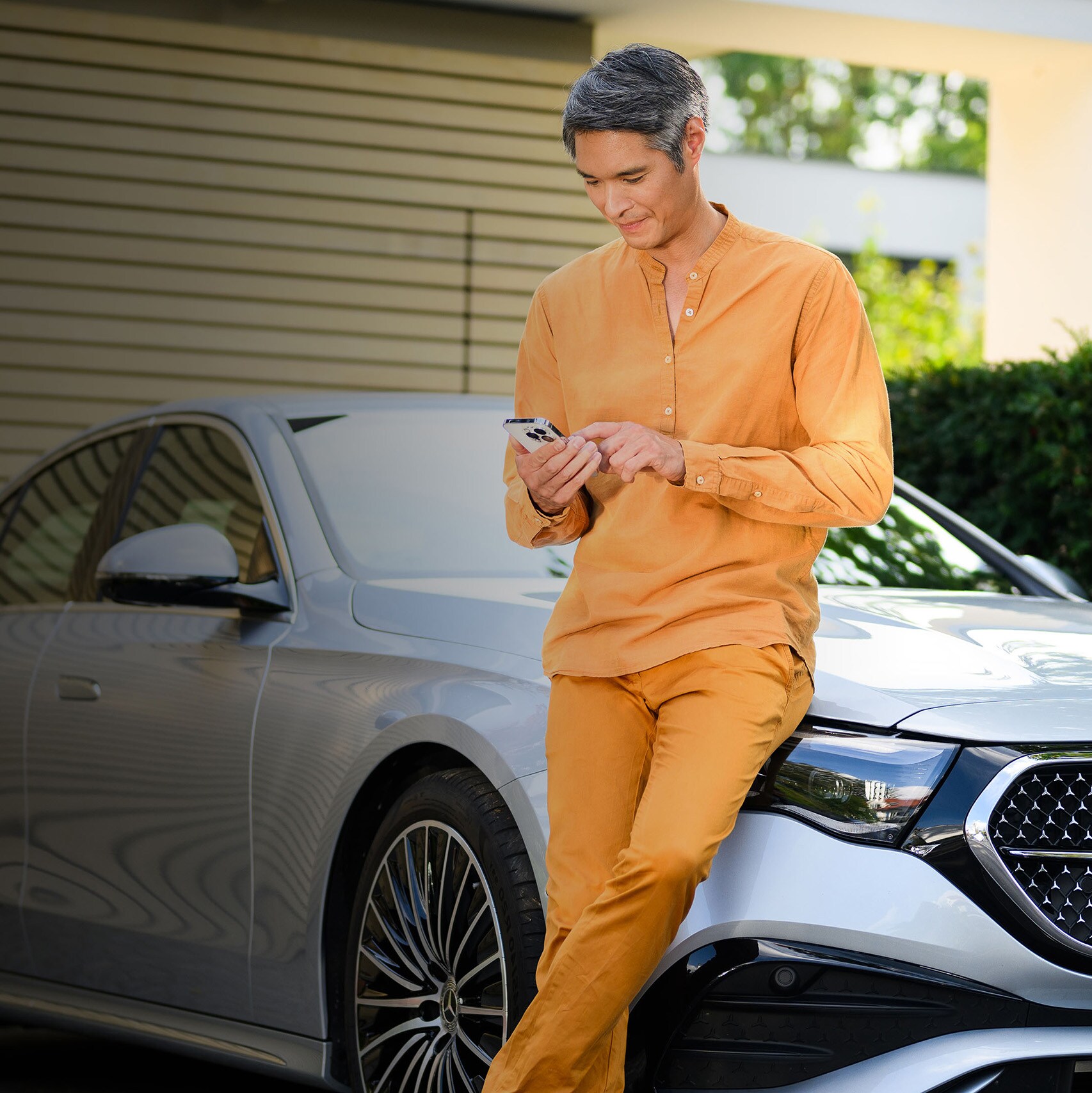 En man i orange kläder använder en mobil framför en silverfärgad Mercedes-Benz.