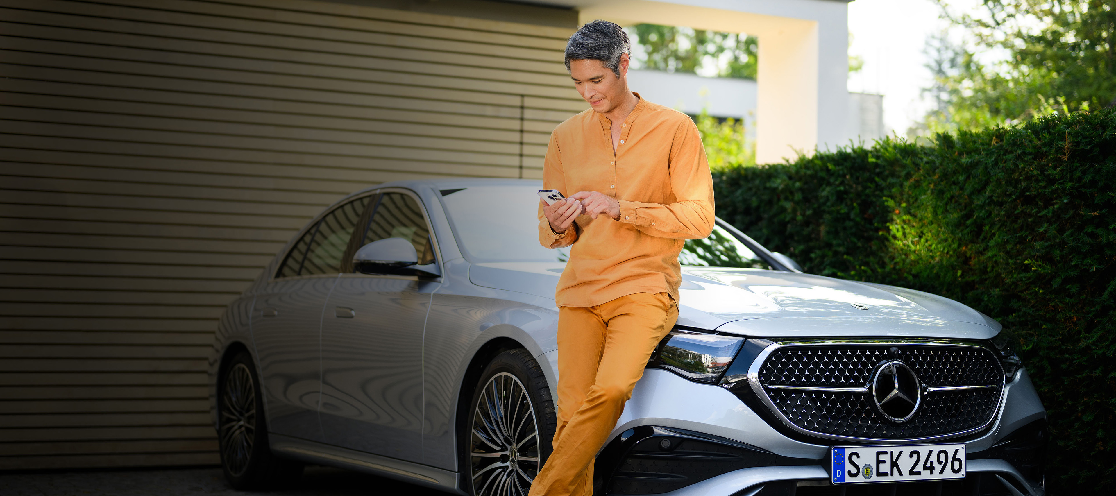 Företagsfinansiering av Mercedes-Benz bilar. En man i orange kläder använder en mobil framför en silverfärgad Mercedes-Benz.