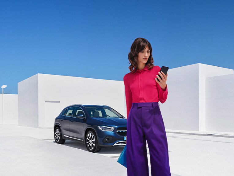 Person utforskar Mercedes-Benz utbud av bilar i sin telefon framför en blå GLA
