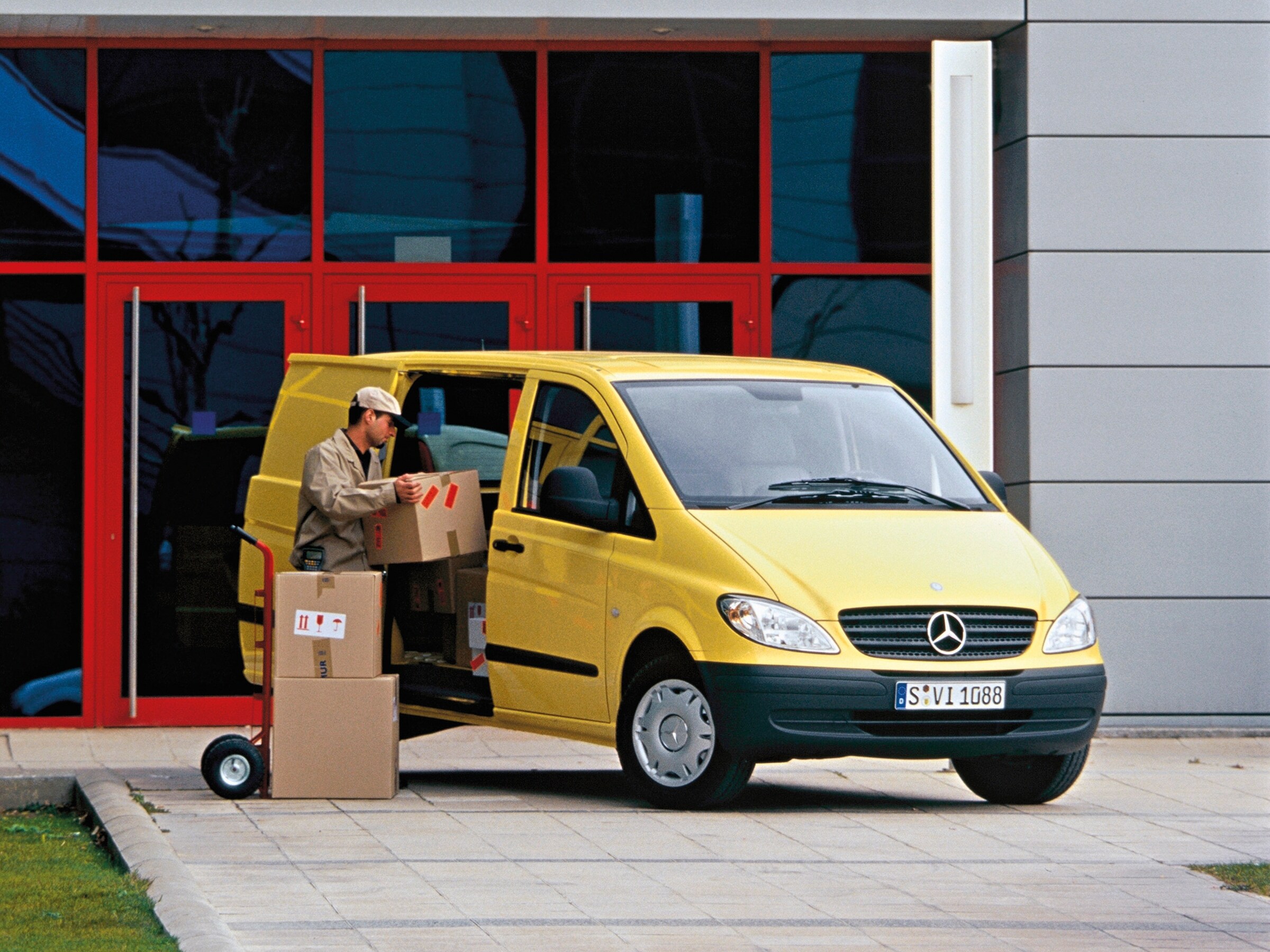 En gul Mercedes‑Benz Vito parkerad utanför en byggnad medan en arbetare lastar av paket