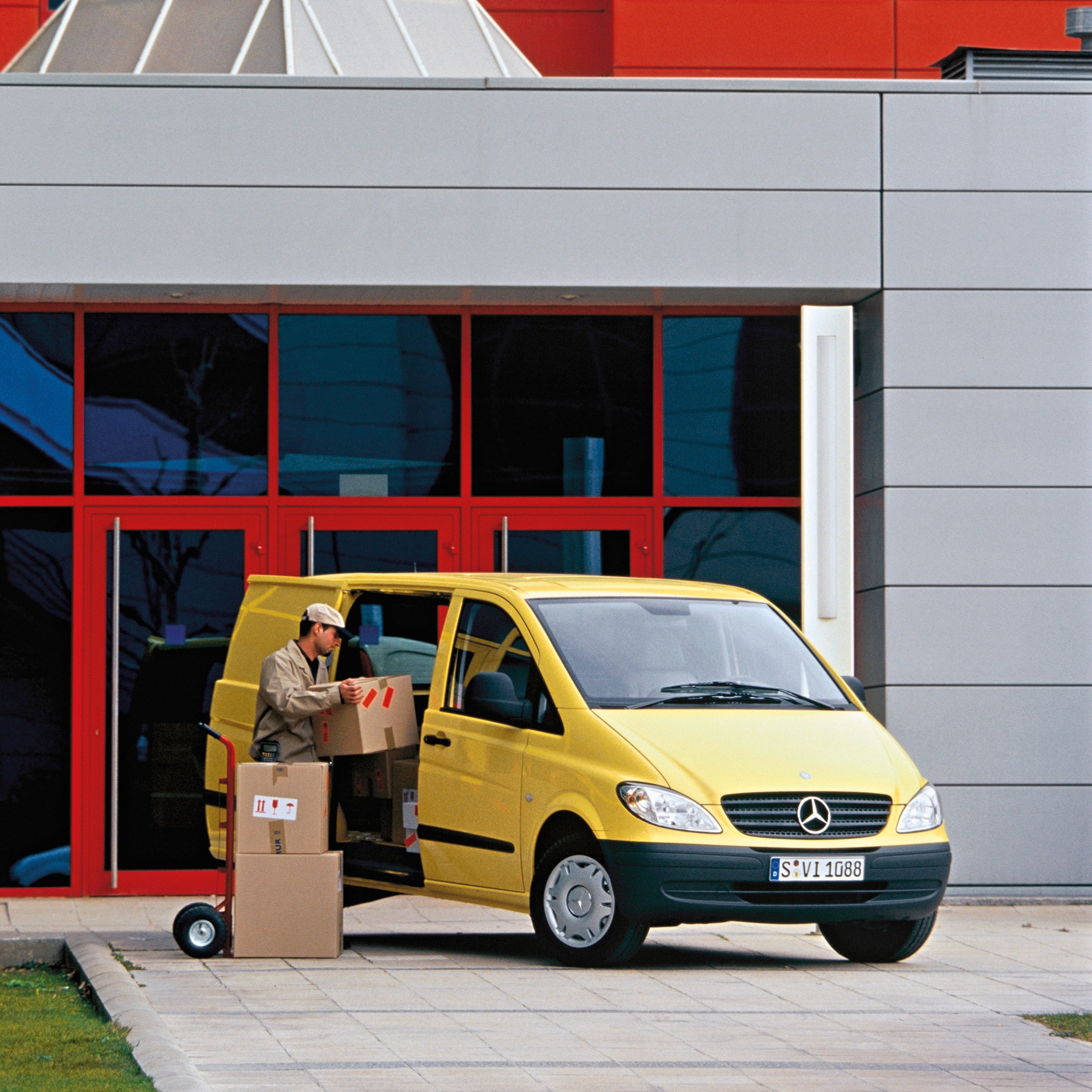 Den andra generationen Mercedes-Benz Vito | Transportbilshistoria | Mercedes-Benz En gul Mercedes‑Benz Vito parkerad utanför en byggnad medan en arbetare lastar av paket