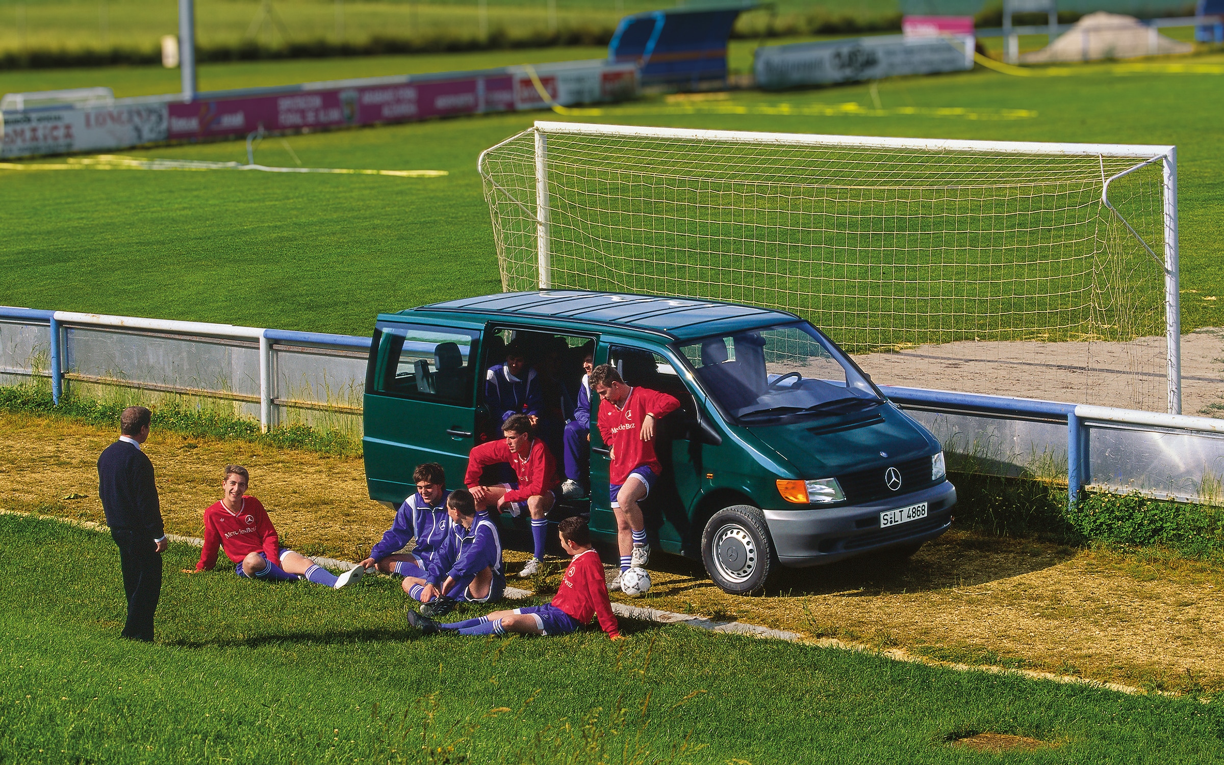 Den första generationen Mercedes-Benz Sprinter | Transportbilshistoria | Mercedes-Benz En Vito i första generationen parkerad vid en fotbollsplan med lagspelare i närheten