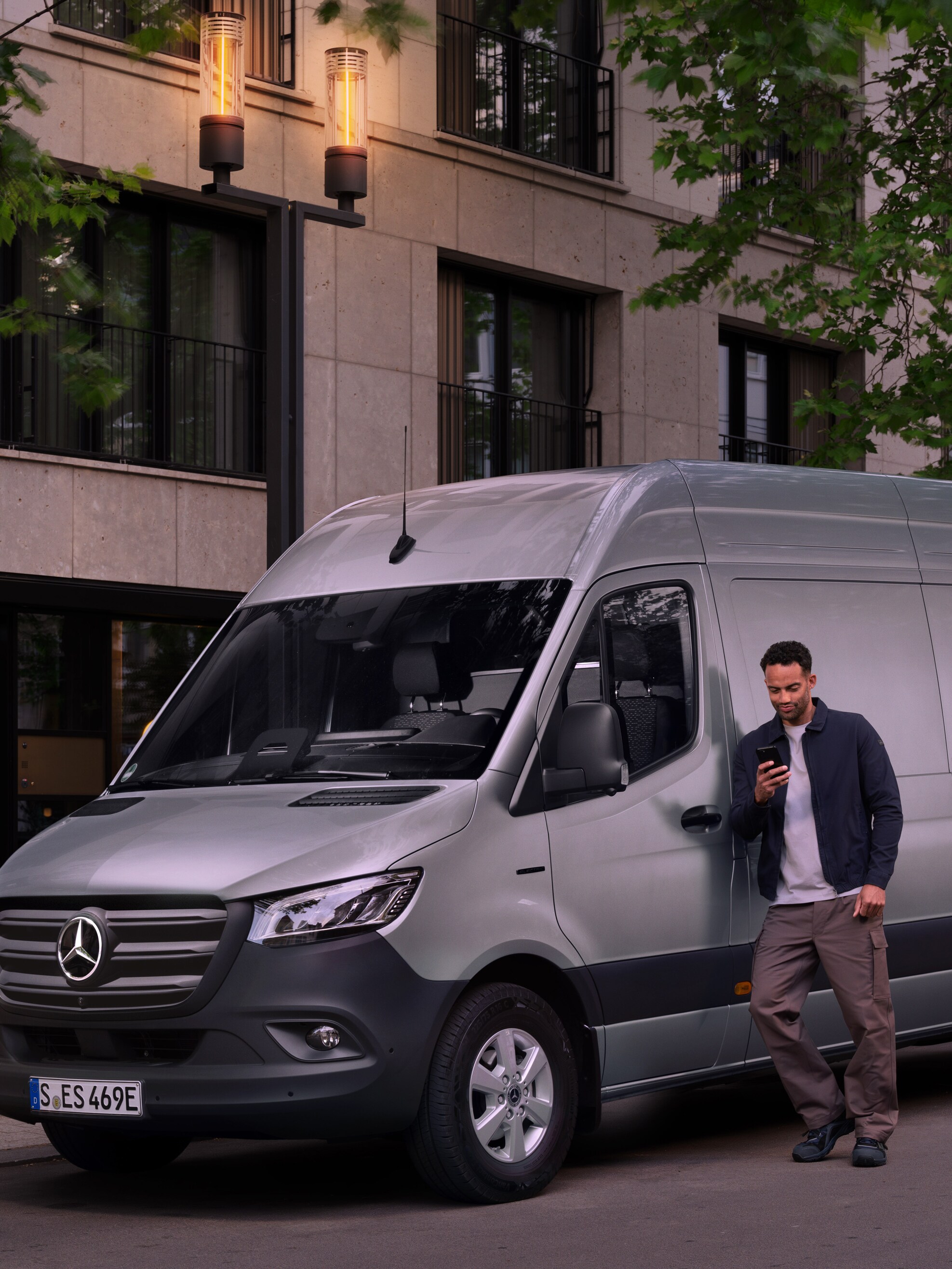 Man lutar sig mot en silverfärgad Mercedes-Benz Sprinter transportbil och använder sin smartphone – snabb hjälp från Mercedes Benz kundtjänst.