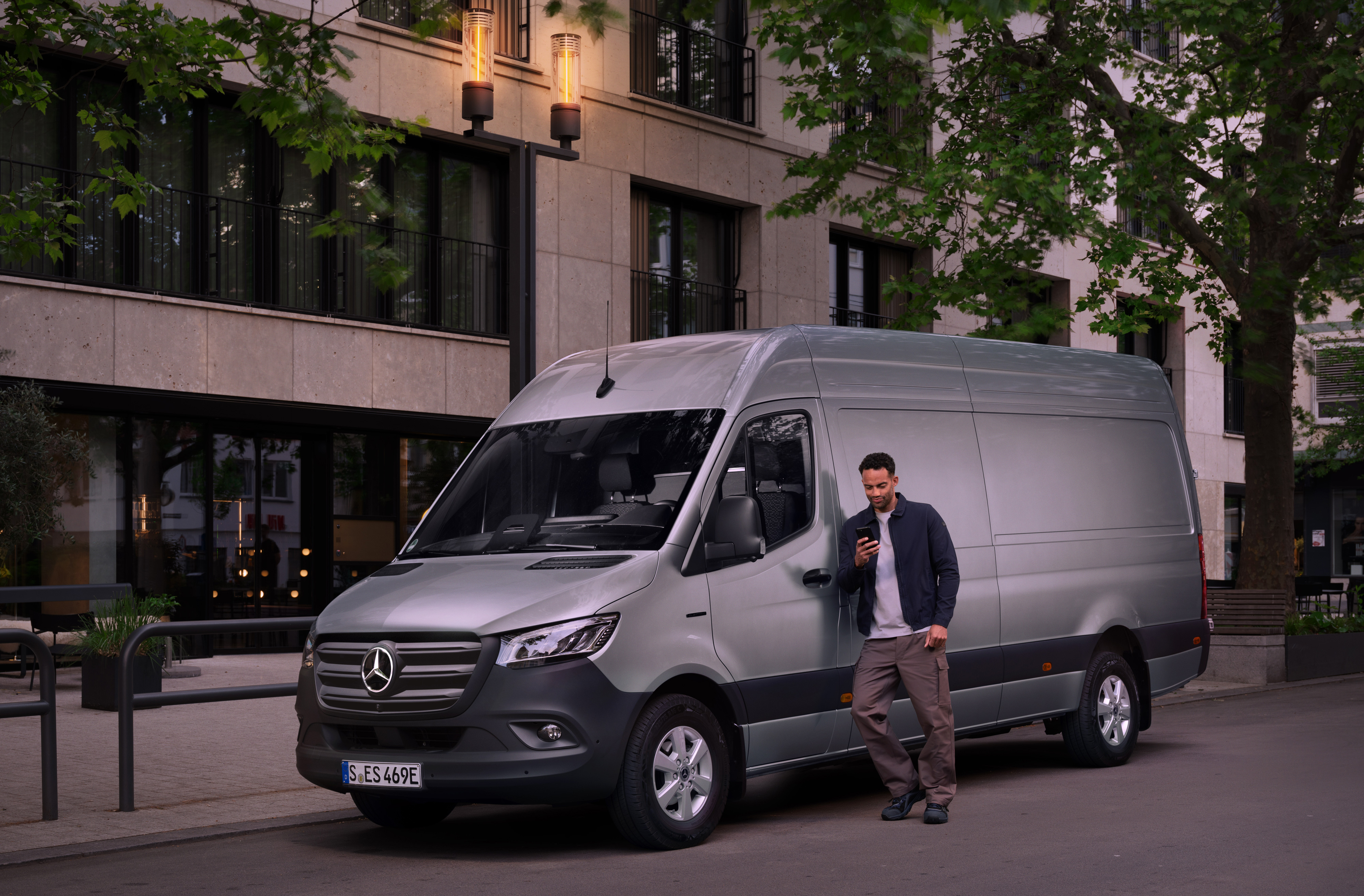 Mercedes-Benz | Van Care | Customer Assistance Center Man lutar sig mot en silverfärgad Mercedes-Benz Sprinter transportbil och använder sin smartphone – snabb hjälp från Mercedes Benz kundtjänst.