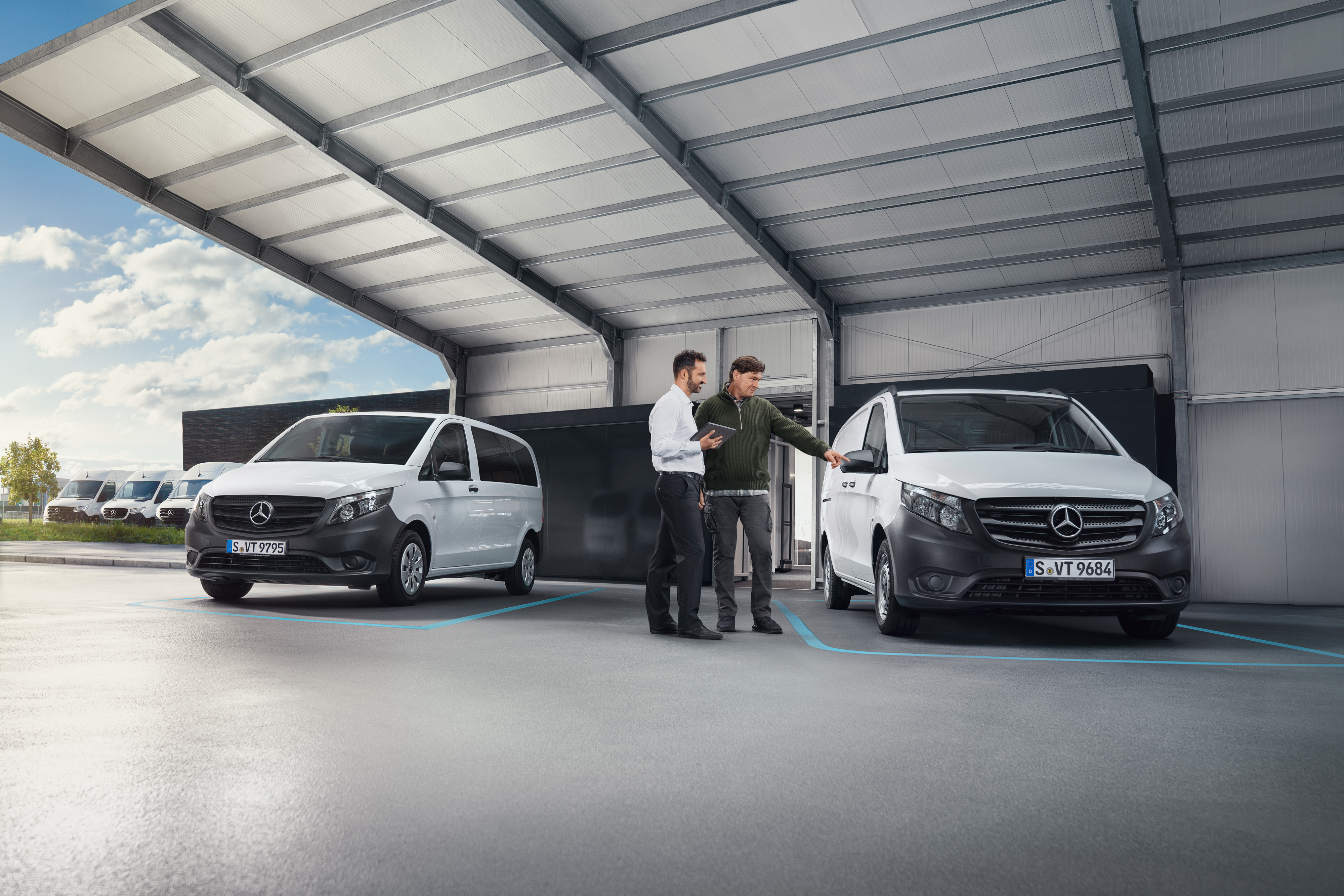 Mercedes-Benz | Bekvämlighet | Digital kundmottagning Två personer pratar i ett arbetsrum framför två Mercedes-Benz-transportbilar.
