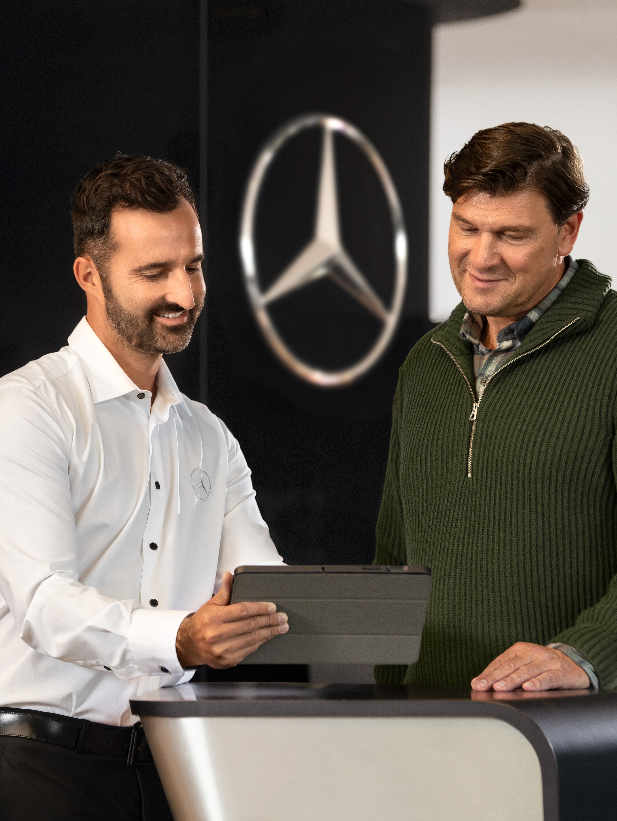 Två män pratar vid en disk samtidigt som de tittar på en surfplatta, framför Mercedes-Benz-stjärnan.