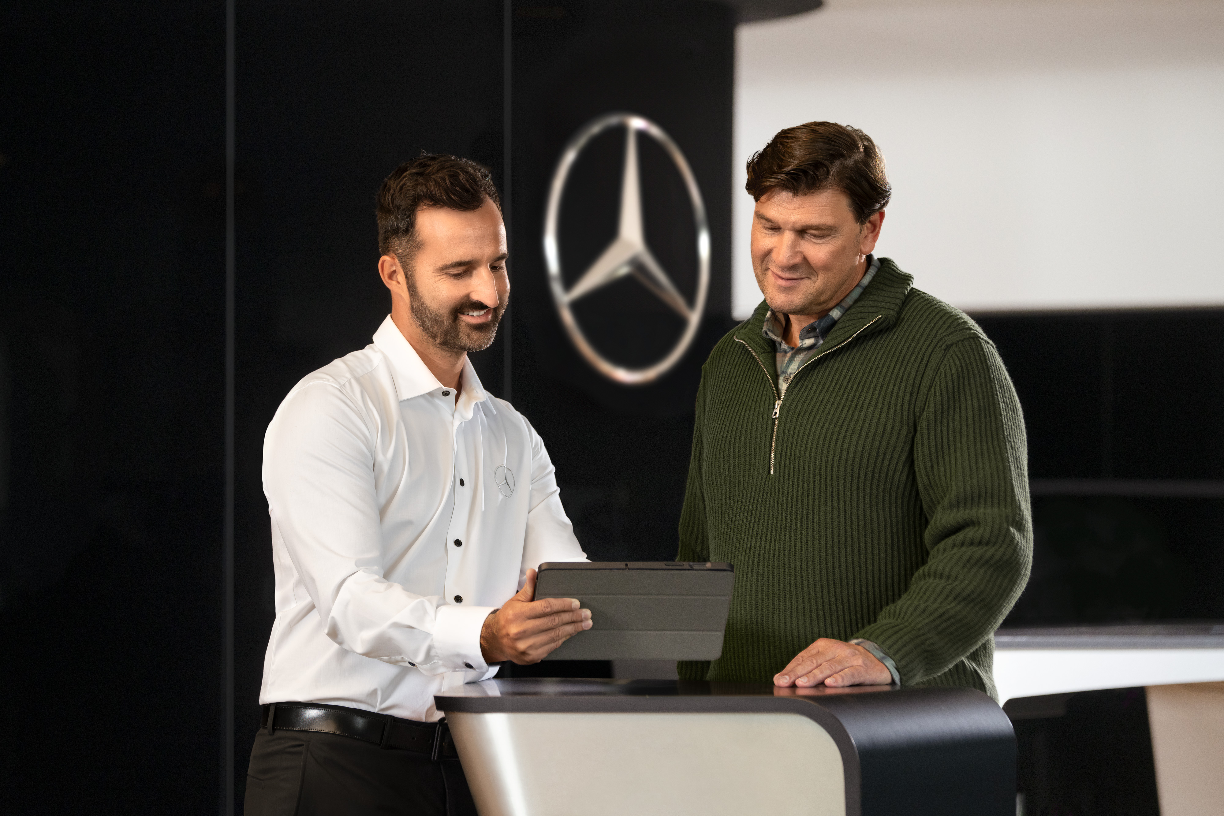 Mercedes-Benz | Översikt över tjänster Två män pratar vid en disk samtidigt som de tittar på en surfplatta, framför Mercedes-Benz-stjärnan.